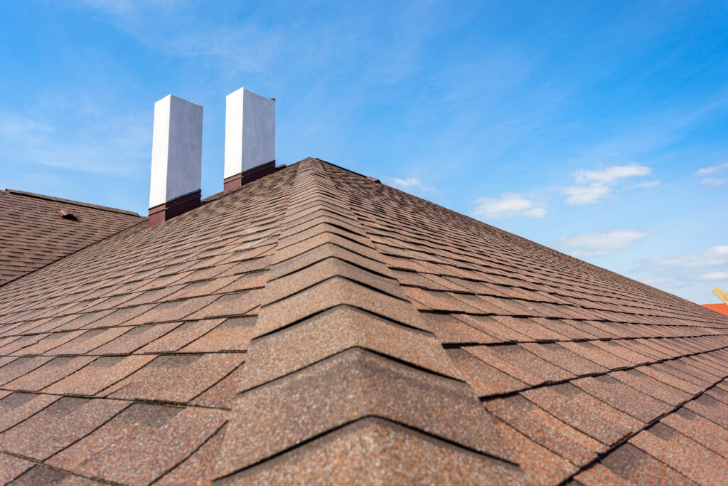 Asphalt shingle roof ridge with chimney stacks under clear blue sky